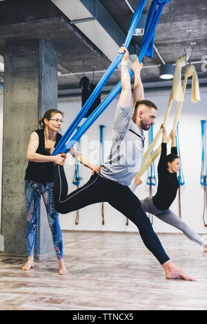Instructor de yoga ayuda a hombre mientras que la mujer ejerza en segundo plano en el gimnasio