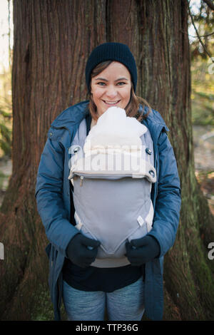 Retrato de la alegre madre con hijo recién nacido en Baby carrier contra el tronco del árbol en el parque Foto de stock