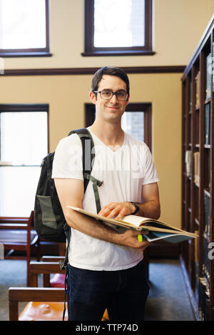 Retrato de seguros estudiante varón con mochila celebración libro mientras está de pie por el estante en la biblioteca