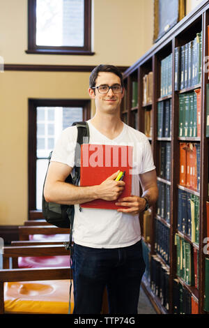 Retrato de seguros estudiante varón con mochila celebración libro mientras está de pie por el estante en la biblioteca