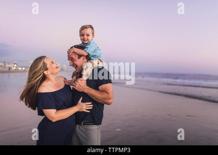Feliz esposo mirando esposa mientras transportaba hijo en la playa contra el cielo durante la puesta de sol