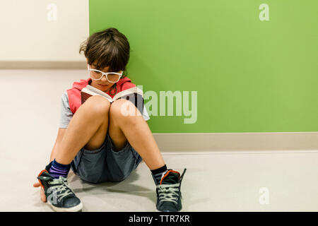 Niño con gafas leyendo sentado en el piso contra la pared inclinada