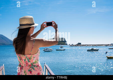 Vista trasera de la mujer fotografiando mar con teléfono inteligente mientras está de pie en el barco contra el cielo azul durante el día soleado