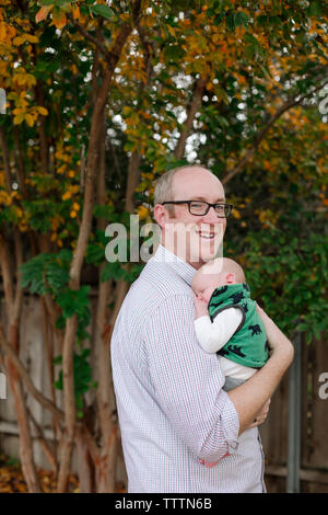 Vista lateral de la sonrisa vertical padre transportando lindo hijo recién mientras está de pie en el parque Foto de stock