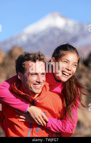 Feliz pareja piggyback en un estilo de vida activo divirtiéndose en la caminata. La joven pareja interracial gozosa. Mujer Asiática caminante y hombre caucásico en actividad al aire libre en el Teide, Tenerife, Islas Canarias, España Foto de stock