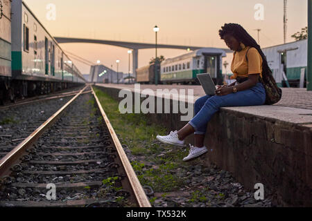Mujer joven sentada en el andén en la estación de tren utilizando el portátil