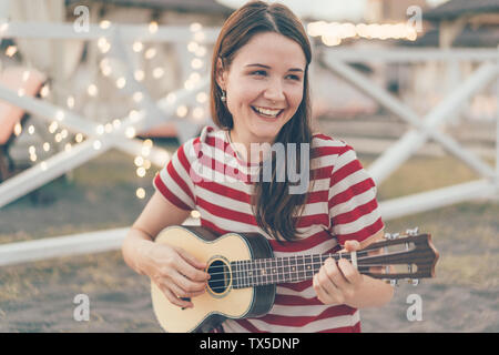 Niña morena jugando un ukulele instrumento acústico y cantar una canción. Festival de música de verano al aire libre. Parte y bokeh garland en el fondo. Foto de stock