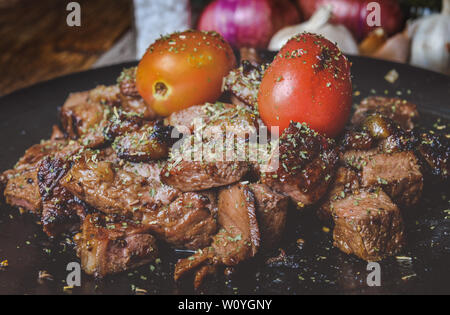 Corte el bistec de carne a la parrilla con especias, condimentos y tomate en la placa negra con grises y tonos oscuros de la iluminación.