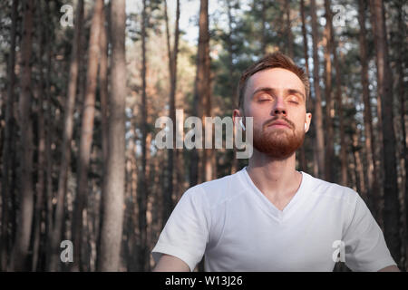 Macho joven persona meditando en el bosque, utilizando tecnología moderna. El hombre se sienta activa en los pinares con los ojos cerrados y disfruta de la meditación aventajar