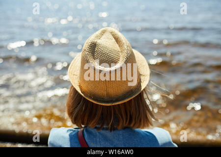Vista posterior retrato de joven mujer vistiendo sombrero de paja mirando al mar en verano, espacio de copia