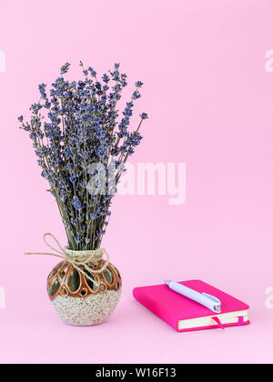 Fragante ramo de lavanda seca con pequeñas flores moradas en un hermoso jarrón de cerámica blanca y bolígrafo en el bloc de notas de papel en una rosa de cubierta Foto de stock