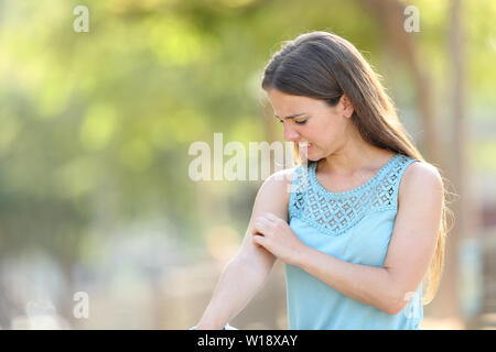 Mujer rascarse el brazo porque las picaduras en un parque con un fondo verde Foto de stock