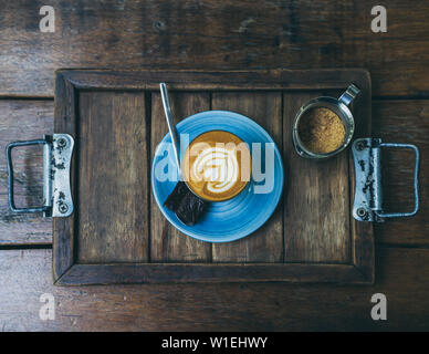 Sentar planas Piccolo Latte café topping con flor de arte de leche en vaso pequeño con un trozo de tarta de brownies caseros en placa de cerámica azul y marrón Foto de stock