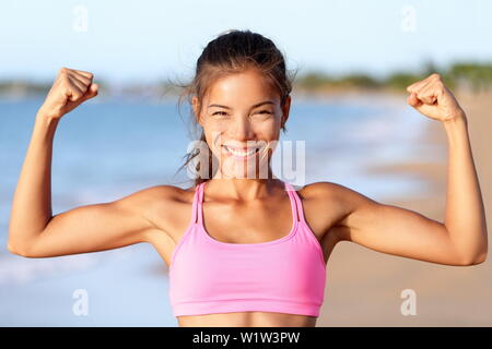 Feliz gimnasia deportiva mujer flexionando los músculos en la playa. Sonriente joven viste rosa sujetador deportivo. Hembra está mostrando su fuerza y estilo de vida saludable en día soleado.