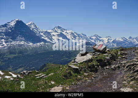 Hiking Trail sign (blanco rojo White Stripes) sobre una roca junto al camino de senderismo con montañas (Eiger, Mönch y Jungfrau) en el fondo. Foto de stock