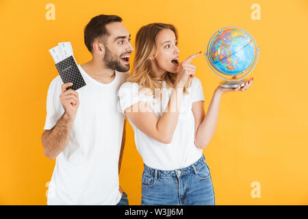Emocionada alegre pareja joven se encuentran aisladas sobre fondo amarillo, sosteniendo globo terrestre, mostrando el pasaporte con billetes de avión Foto de stock