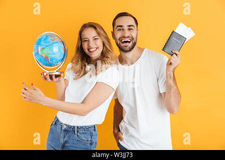 Emocionada alegre pareja joven se encuentran aisladas sobre fondo amarillo, sosteniendo globo terrestre, mostrando el pasaporte con billetes de avión Foto de stock