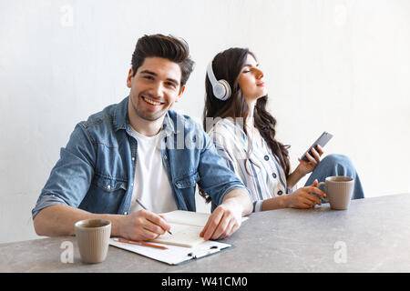 Atractiva pareja joven bebiendo café en la mañana en casa, sentado a la mesa, estudiar, escuchar música con auriculares Foto de stock