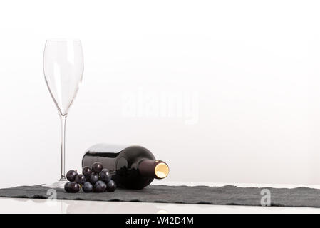 La viticultura y el concepto de vino con un elegante wineglass vacía junto a un racimo de uvas negras y sin etiquetar, botella de vino tinto a su lado contra un Foto de stock