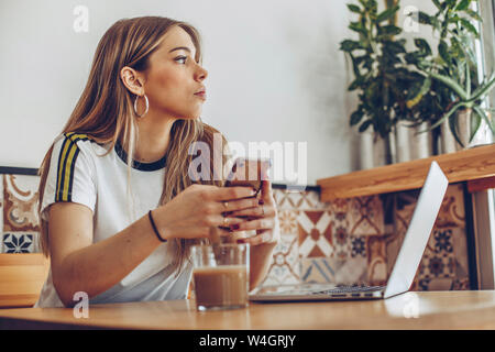 Mujer joven con el teléfono móvil y el ordenador portátil en la cafetería