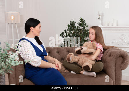 Madre e hija pasan tiempo juntos, sentarse en el sofá y chat. Ocio madres e hijas. Niña triste abrazos Teddy Vedmed y llora cerca de mamá. Foto de stock