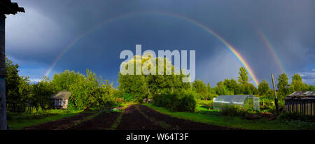 Panorama de verano con un doble arco iris en el cielo espectacular por encima de la aldea cocina-jardín con un invernadero y casa pequeña Foto de stock