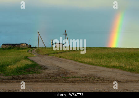 Doble arco iris en el cielo a través de una pequeña granja con postes de electricidad y un camino entre prados verdes Foto de stock