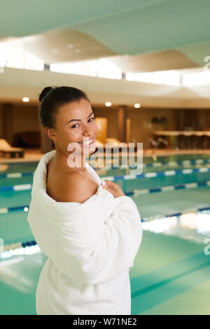 Retrato de hermosa raza mixta mujer vistiendo albornoz de pie junto a la piscina en el balneario y mirando a la cámara, espacio de copia Foto de stock