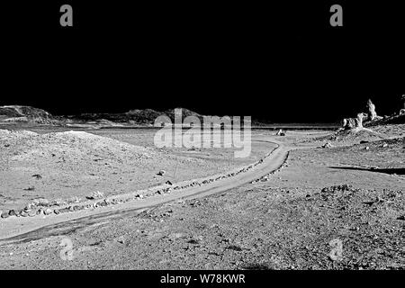 Camino de tierra curvándose a través del árido desierto valle con colinas y formaciones rocosas bajo el negro cielo. Blanco y negro. Foto de stock