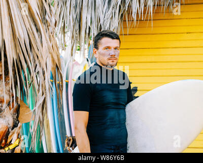 Retrato de un surfista en la cara de un protector solar, vistiendo pantalones cortos y una camiseta negra de pie con una tabla de surf. amarillo fondo de madera.
