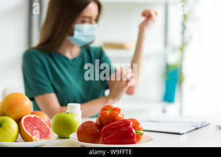 El enfoque selectivo de la mujer joven en máscara de médico sentado a la mesa con las frutas, las verduras y las píldoras y rascarse el brazo en casa Foto de stock