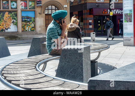 Un sij hombre se sienta en un banco en la calle High Street en el centro de la ciudad de Slough y habla en su teléfono móvil Foto de stock