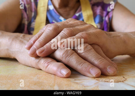 Cerca de las manos de una mujer mayor juntos en una mesa de la cocina. La vieja señora vistiendo coloridos pinafore, el enfoque selectivo, iluminación natural, vista delantera. Foto de stock
