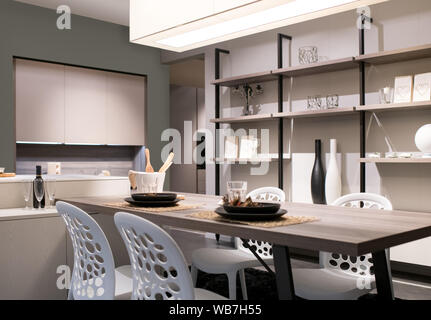 Cocina y comedor con decoración beige neutro, Estanterías de pared y una moderna unidad de sillas y mesa iluminada por una gran sobrecarga de luz de techo Foto de stock