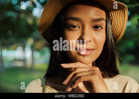 Retrato de mujer vistiendo lindo piercing labial y sombrero de paja tocando su barbilla mientras mirando a la cámara en el parque verde Foto de stock