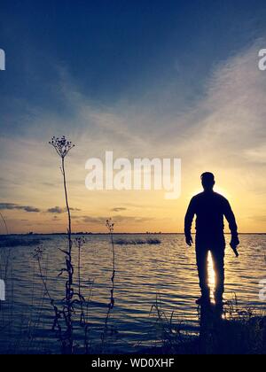 Silueta el hombre con la pistola de pie sobre una roca en el lago contra el cielo durante la puesta de sol