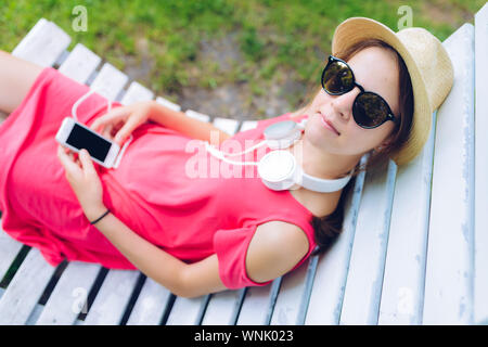 Joven descansando sobre una silla de madera colgadas en el jardín - con auriculares alrededor de su cuello y un smartphone en las manos Foto de stock