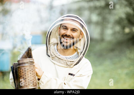 Retrato de un alegre apicultor en uniformes de protección de abejas en el apiario fumador Foto de stock