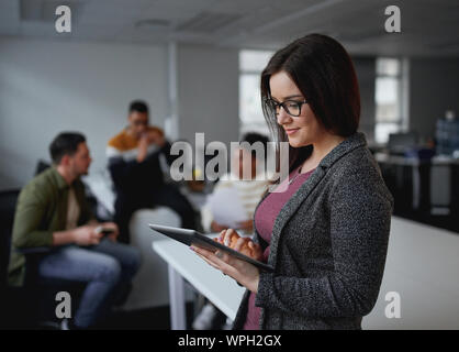 Feliz joven empresaria mediante tableta digital en la oficina y sus colegas discutiendo en segundo plano.