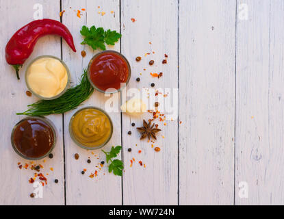 Un conjunto de salsas y uno especial: ketchup, mayonesa y mostaza en una placa blanca, pimiento picante, perejil, eneldo. Vista superior en fondo blanco de madera. Foto de stock