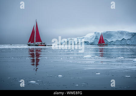 Hermoso color rojo velero en el ártico junto a un enorme iceberg que muestra la escala. Navegando entre témpanos flotantes en la bahía Disko glaciar durante Foto de stock