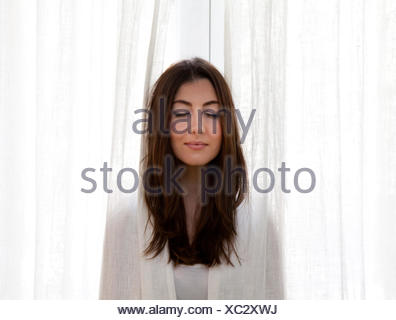 Adulto joven mujer metiendo la cabeza de cortinas, los ojos cerrados