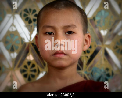 Bouddhiste birman sérieux garçon moine au pagode Su Taung Pyae pose pour une photo officielle devant un miroir-modelé (Tiens-réalisatrice) temple mur. Banque D'Images