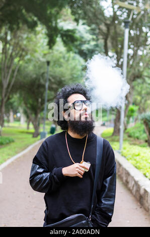 Vape. Jeune homme brutal avec grande barbe et lunettes à la mode coupe de cheveux dans un tabac cigarette électronique dans le parc de la ville. Nuage de vapeur. Style de vie. Banque D'Images