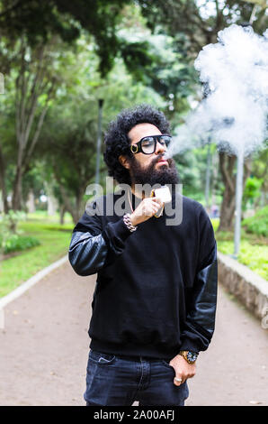 Vape. Jeune homme brutal avec grande barbe et lunettes à la mode coupe de cheveux dans un tabac cigarette électronique dans le parc de la ville. Nuage de vapeur. Style de vie. Banque D'Images