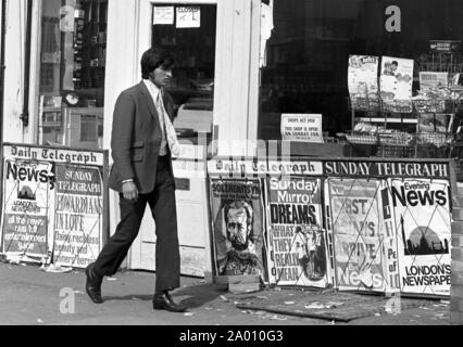 Southall, Londres 1972. Un design habillé jeune indien homme en passant devant un marchand de journaux avec un 'Premier annonces indiquant les asiatiques arrivent..'. Après Idi Amin commandé 30 000 Asiatiques hors de l'Ouganda avec préavis de 90 jours, ceux avec un passeport britannique dirigé vers l'Angleterre. Ils ont été forcés de quitter leurs fonds, leurs biens et possessions derrière. Ces images documentent la vie de certaines des personnes lorsqu'ils se sont installés dans la région de Southall, à l'ouest de Londres. Un aperçu de la vie il y a 47 ans pour les immigrés au Royaume-Uni. Photo par Tony Henshaw Banque D'Images
