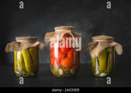 Les légumes en conserve de tomates et de concombre dans des bocaux en verre noir sur planche de bois. Vue de côté. Récolte et préparations maison de l'automne. Banque D'Images