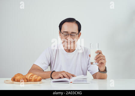 Vieil homme boire du lait et de la lecture livre tout en ayant le petit déjeuner Banque D'Images