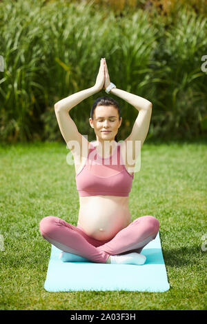 Young pregnant woman sitting in lotus position sur tapis d'exercice a soulevé ses bras et faisant exercice de relaxation dans le parc Banque D'Images