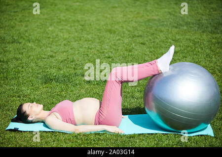 Young pregnant woman lying on exercise mat a élevé ses pieds sur fitness ball et de détente avec ses yeux fermés à l'extérieur Banque D'Images